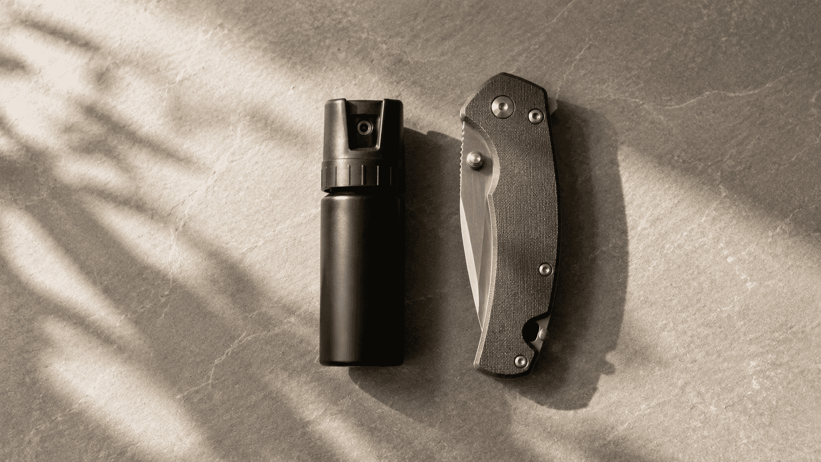 Flat lay editorial photograph showing a compact pepper spray canister and a closed folding knife arranged side by side on a neutral stone surface with balanced spacing Soft natural window light creates gentle shadows across muted earth tones producing a calm and thoughtful product comparison style scene that conveys careful consideration and responsible preparedness
