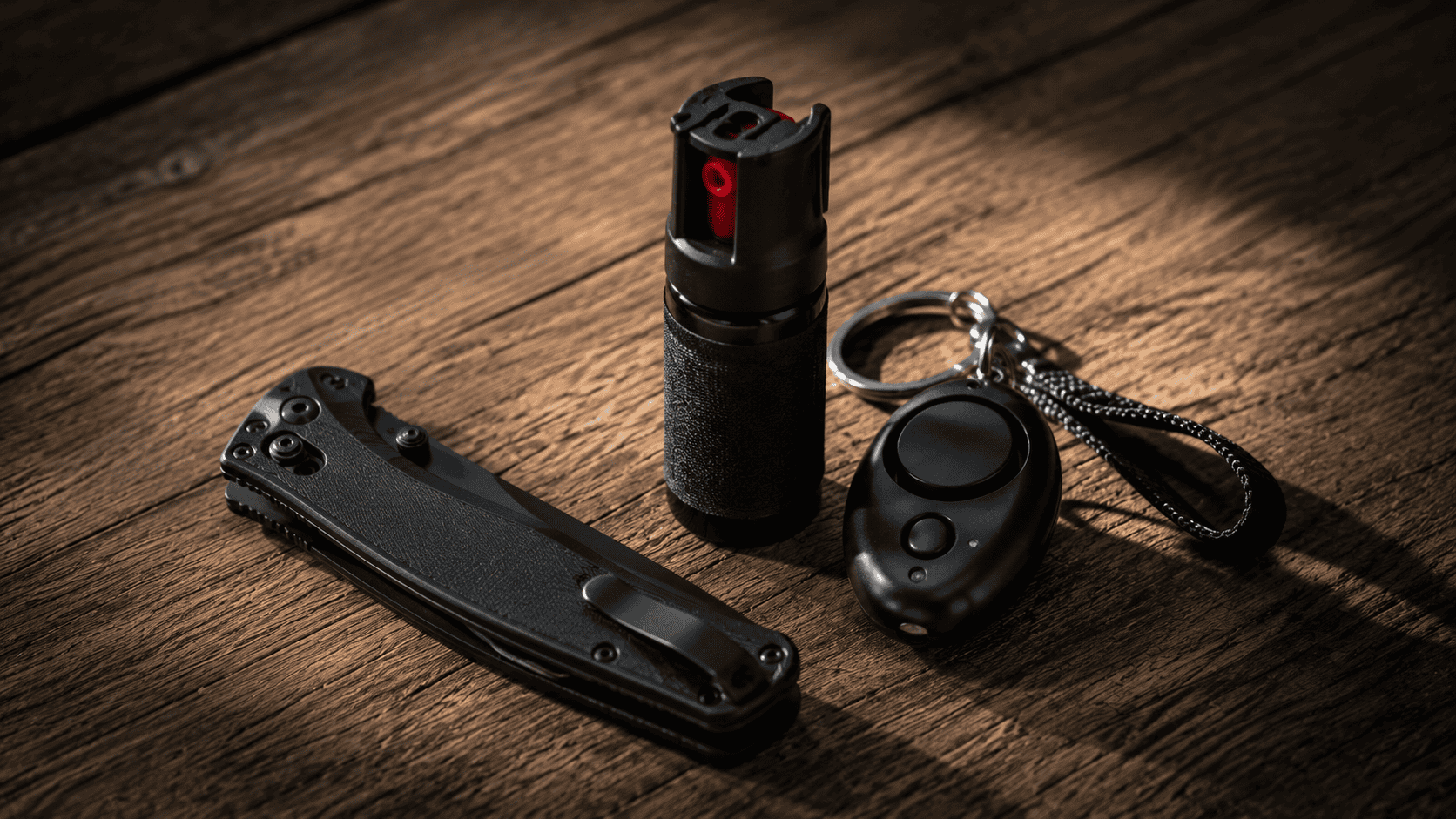 Closed black folding pocket knife beside pepper spray and a keychain personal alarm on a wooden surface